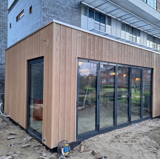 Foto van de nieuwe tuinkamer in aanbouw in de binnentuin van Ter Reede. Bekleed met houten latten en zwarte kozijnen.